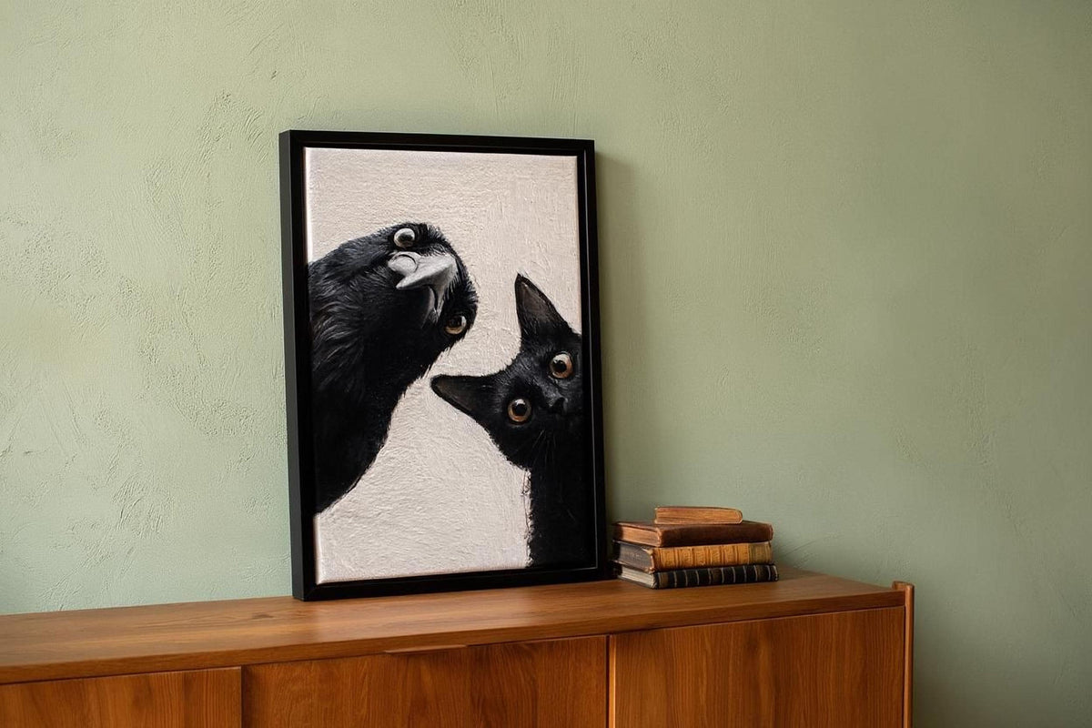A framed painting of a black cat peeking out from behind a bird is displayed on a wooden cabinet against a green wall.
