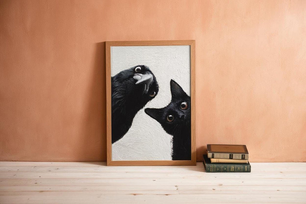 A framed photograph of two black cats peeking out from behind a wooden frame is displayed on a wooden floor against a peach-colored wall.
