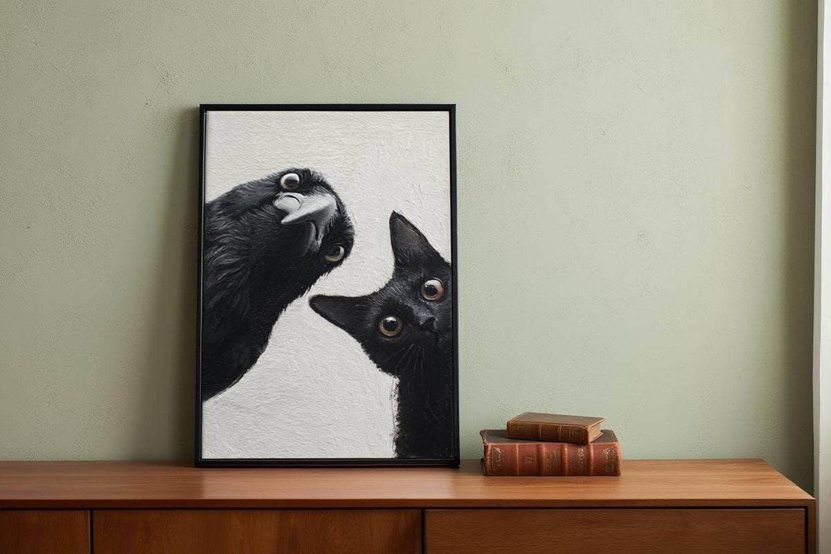 A framed photograph of a black cat peeking out from behind a black bird is displayed on a wooden dresser next to a stack of books.
