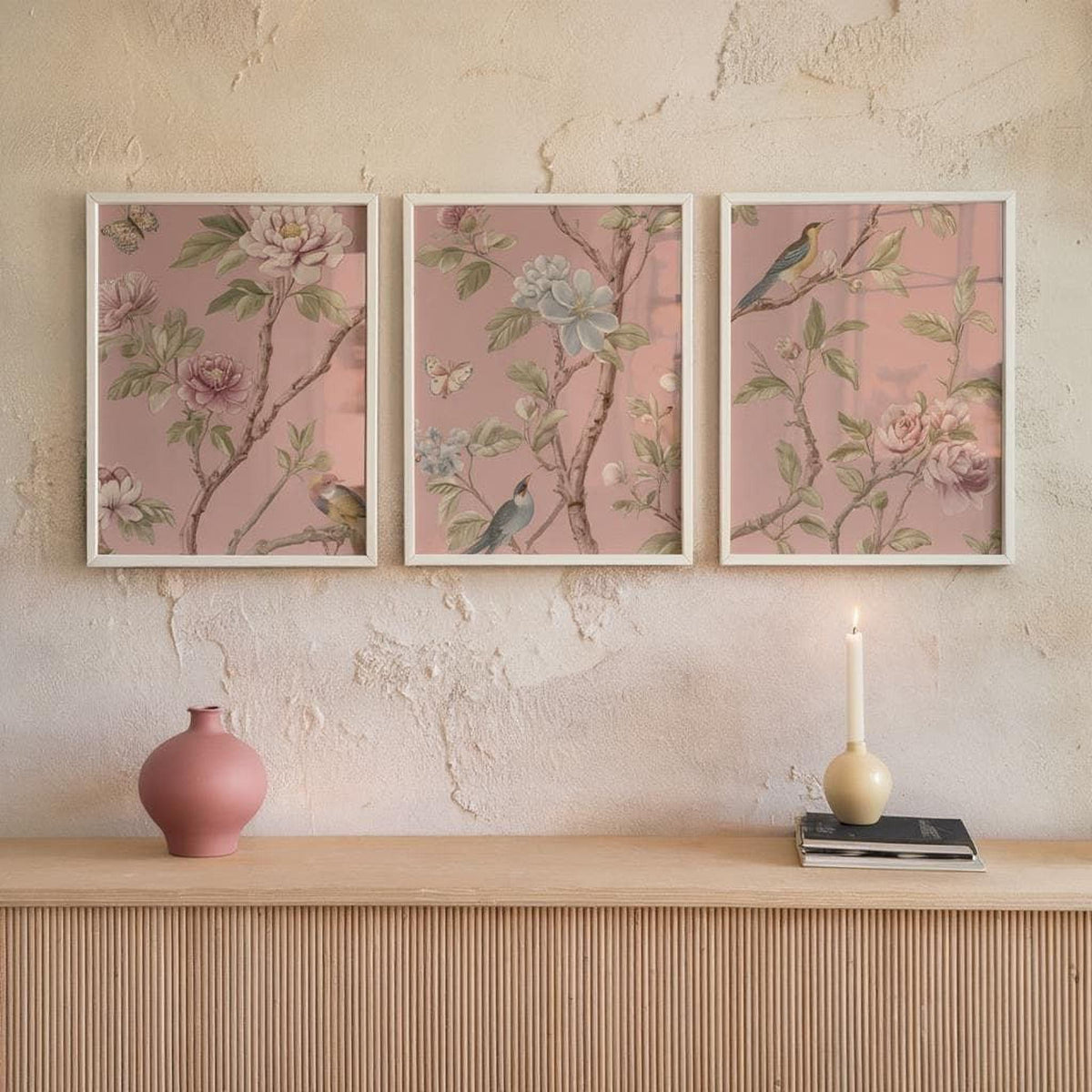 a shelf with three framed pictures of flowers hanging on a wall above it. There is also a pink vase and a lit candle on the shelf.