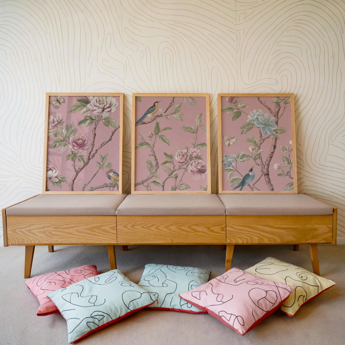 a wooden bench with three framed floral designs on the backrest, arranged in a row. The bench is placed on a carpeted floor, and there are several colorful throw pillows on the ground in front of it.