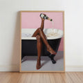 Framed print leaning on wooden floor, black woman bathroom wall art, legs crossed pouring champagne into clawfoot tub