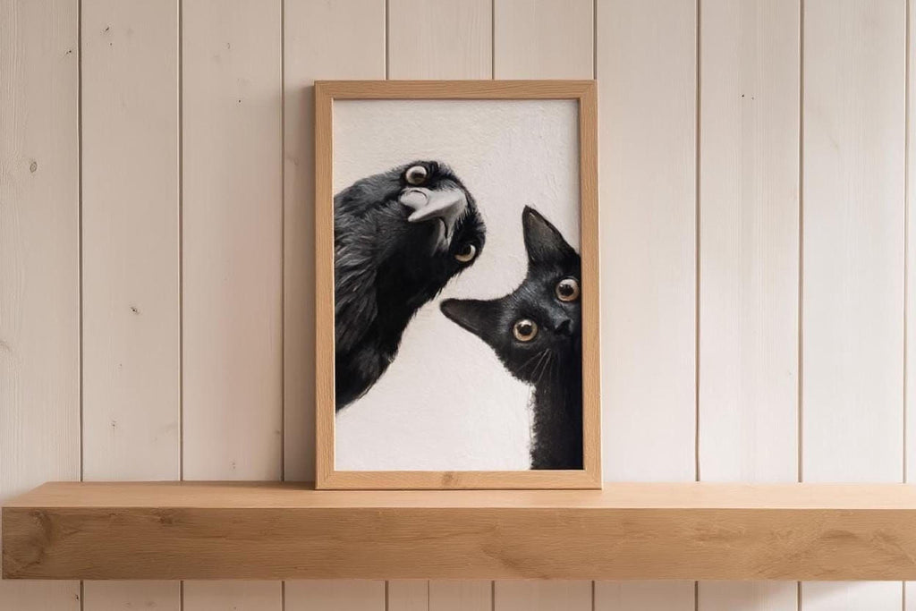 A framed image of a black cat peeking out from behind a black bird, both with wide-open eyes, is displayed on a wooden shelf against a white wooden wall.
