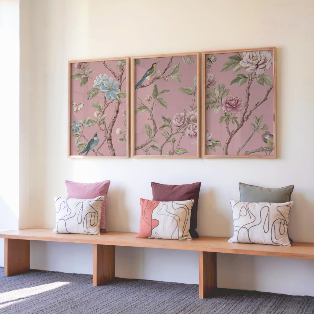 a cozy seating area with a wooden bench and three decorative pillows, each with a unique design. Above the bench, there are three framed pictures hanging on the wall, depicting a pink floral pattern.