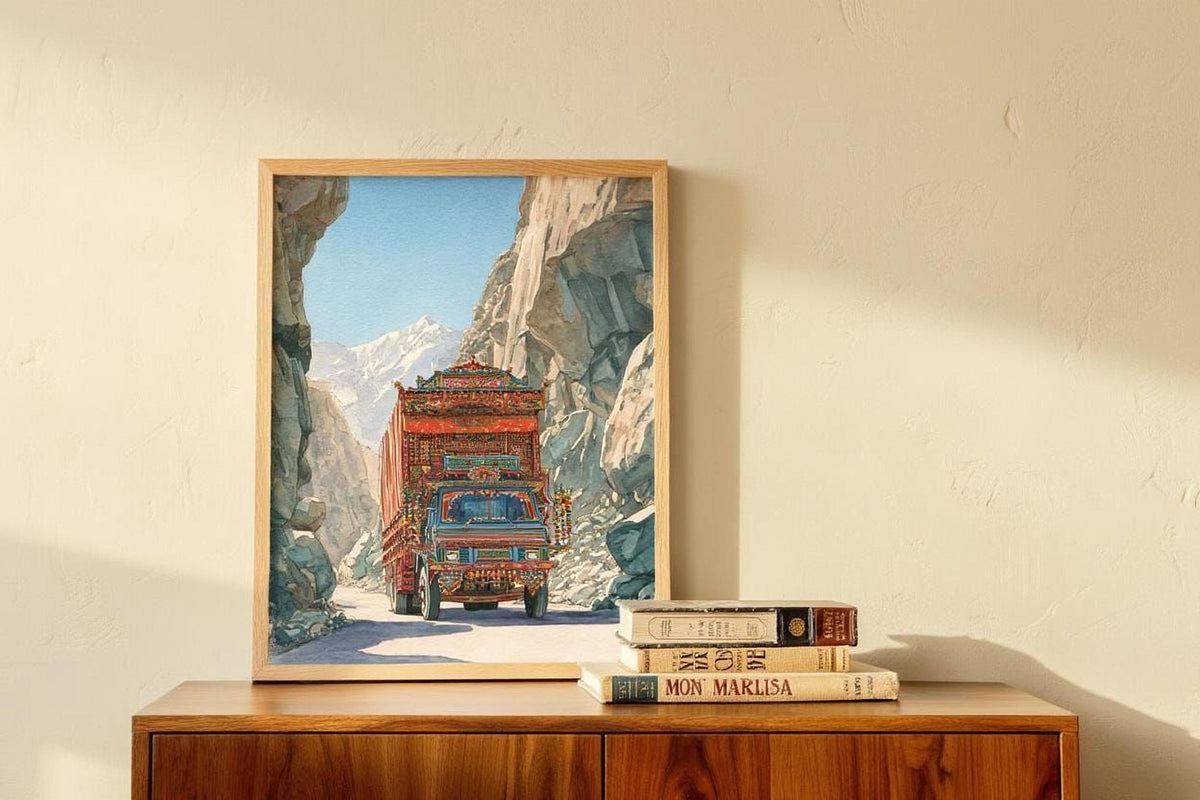 A framed painting of a colorful truck driving through a mountainous landscape is displayed on a wooden cabinet, accompanied by a stack of books.