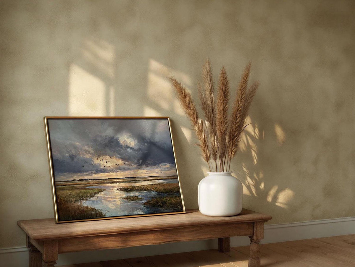 a wooden table with a framed landscape painting and a vase of dried reeds placed on it. The painting depicts a serene landscape with a body of water and a cloudy sky. The vase contains dried reeds, adding a natural element to the scene. The table is positioned against a beige wall, and the lighting in the room creates a warm and inviting atmosphere.