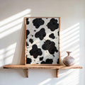 Framed cow print wall art leaning on a floating wood shelf, sunlit window stripes, small pottery