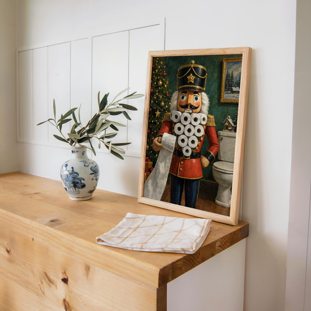 Framed funny nutcracker bathroom print on a shelf with a blue vase and folded linen, versatile holiday wall decor.