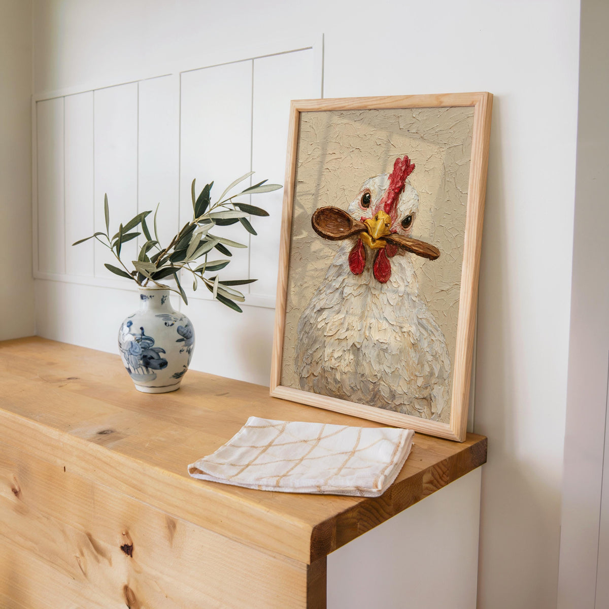 Leaned hen holding spoon print on a farmhouse desk beside blue vase and olive sprig, chicken kitchen wall art