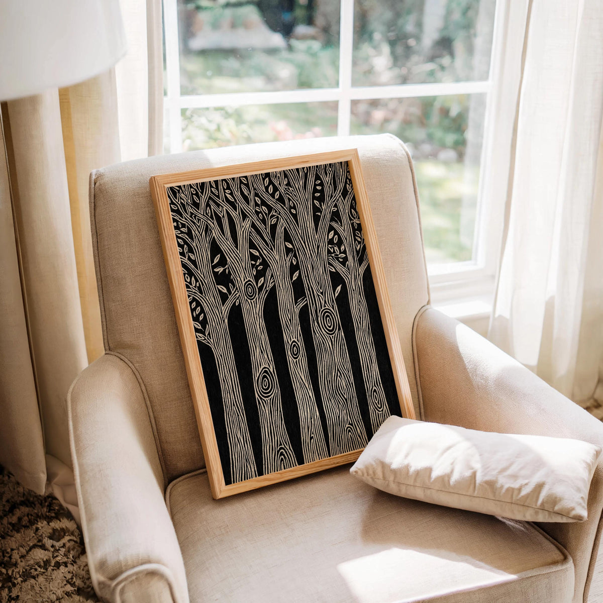 Framed black and white forest wall art leaning on a beige armchair beside a bright window, monochrome nature print for cozy rooms