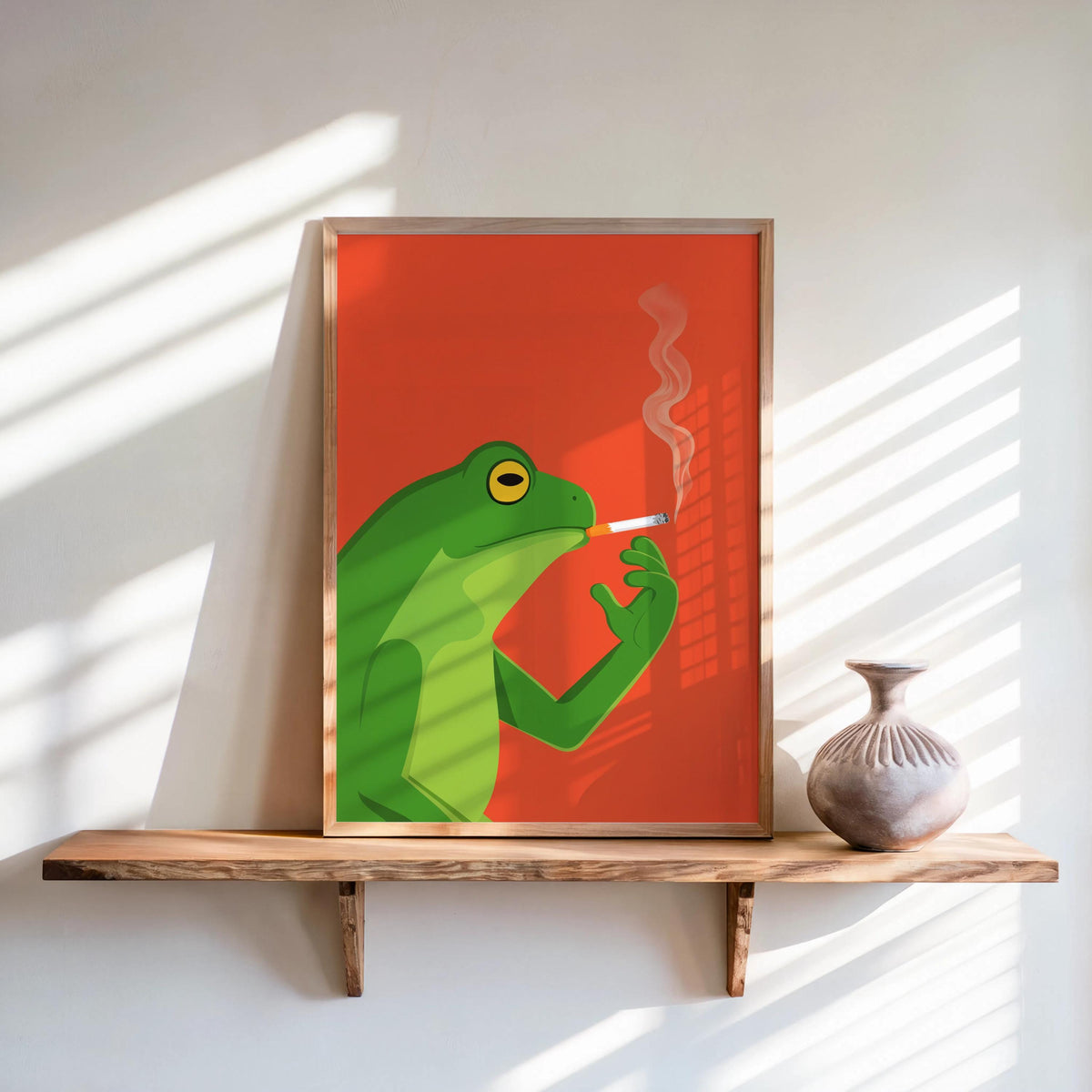 Framed funny frog wall art print leaning on sunlit wood shelf, bold red-orange retro vibe