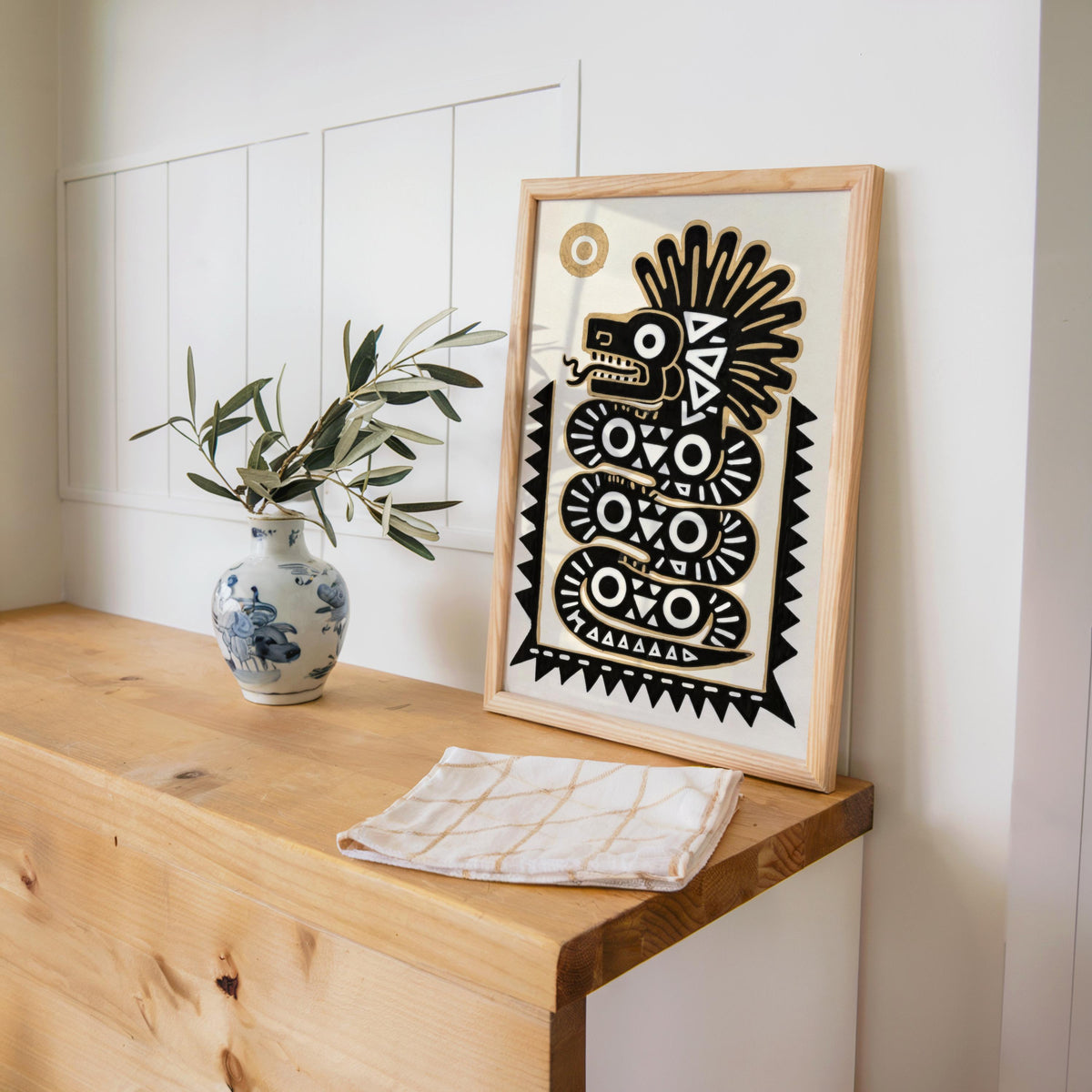 Framed indigenous mexican art on a corner shelf beside a blue ceramic vase and leafy plant, soft neutral tones