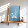 Framed blue romantic swan wall art on wooden shelf, two white swans forming a heart, textured minimalist print