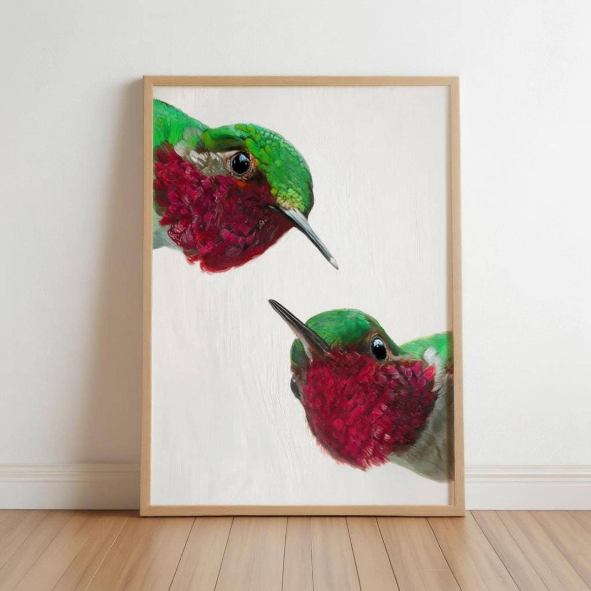 Light-wood framed whimsical hummingbird wall art print leaning on white wall, vivid red and green birds.