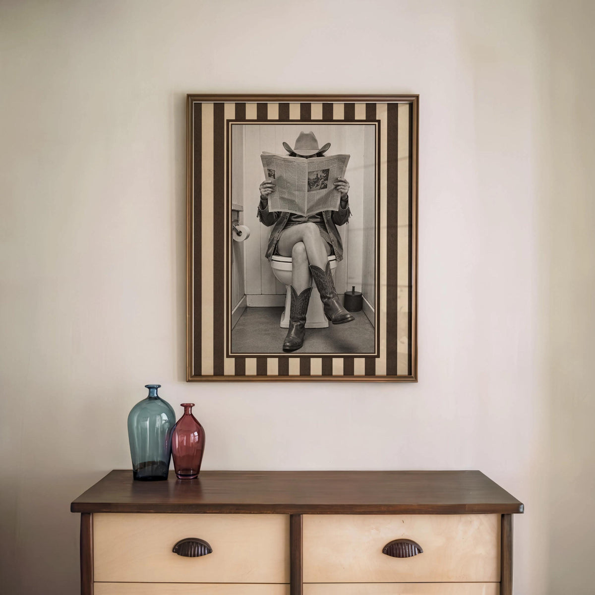 Vertical striped-border cowgirl print centered above a wood console with two vases, funny cowgirl bathroom art, vintage cowgirl print