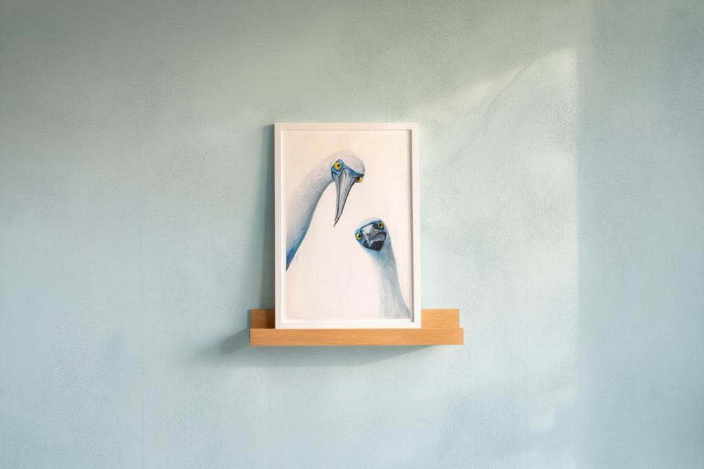 Small framed print on narrow wooden shelf against cool teal wall, coastal charm and quirky character, blue footed booby wall art print