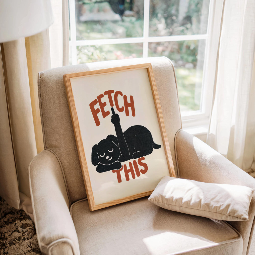 Framed print nestled on a cushioned armchair, charcoal dog silhouette and rust-red FETCH THIS type, rude dog wall decor, funny dog wall art