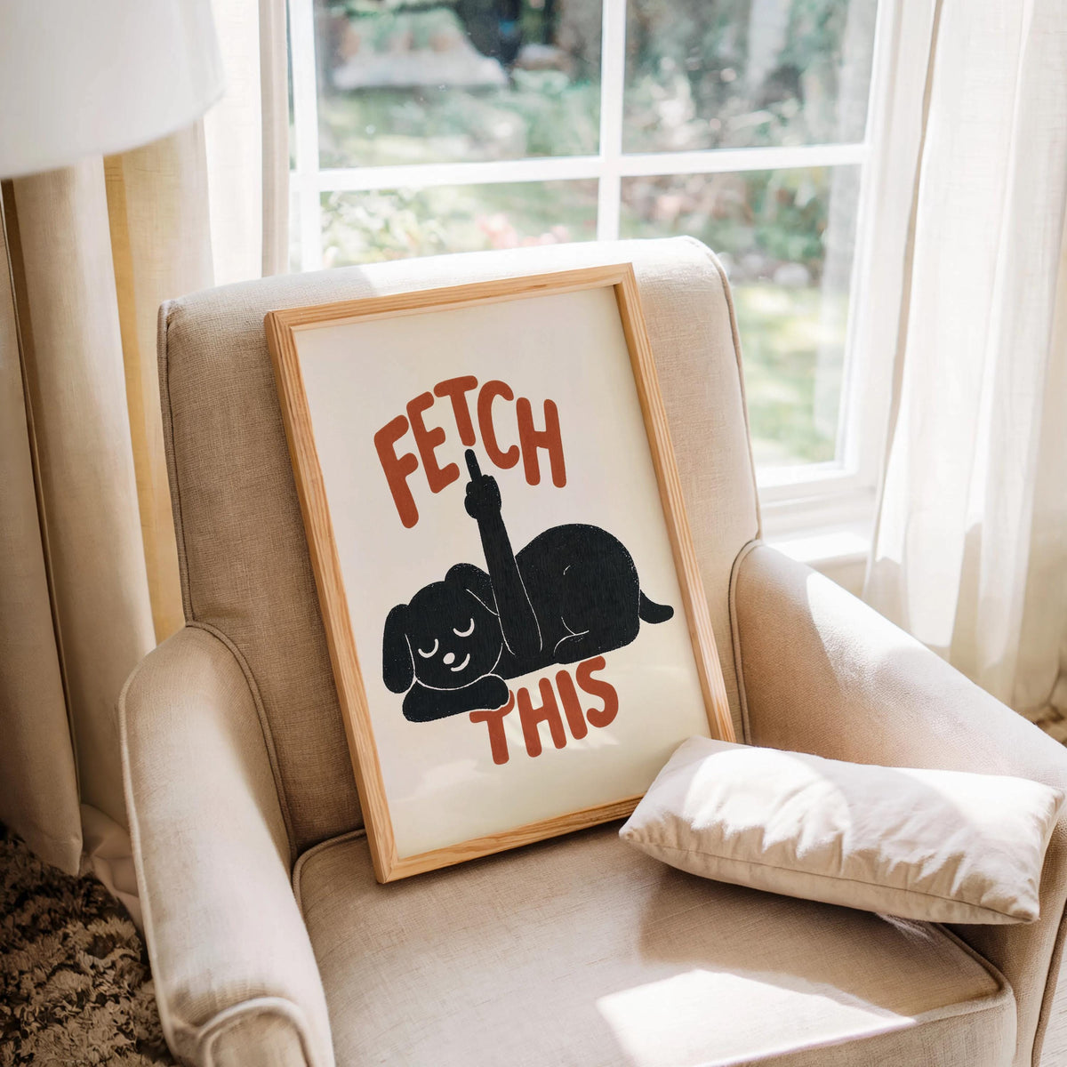 Framed print nestled on a cushioned armchair, charcoal dog silhouette and rust-red FETCH THIS type, rude dog wall decor, funny dog wall art