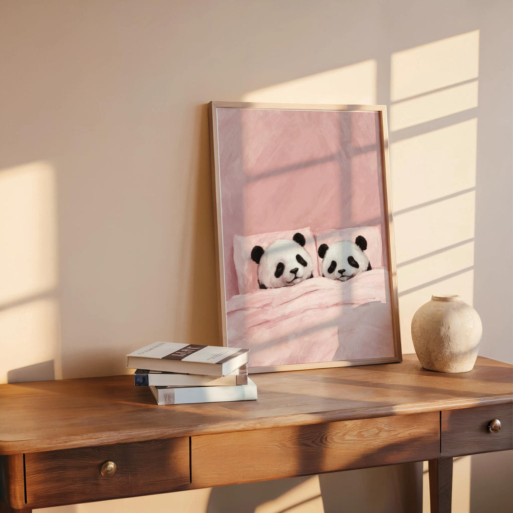 Blush sleeping panda wall art leaning on a wooden desk, stacked books and round vase, cozy nursery print vibe