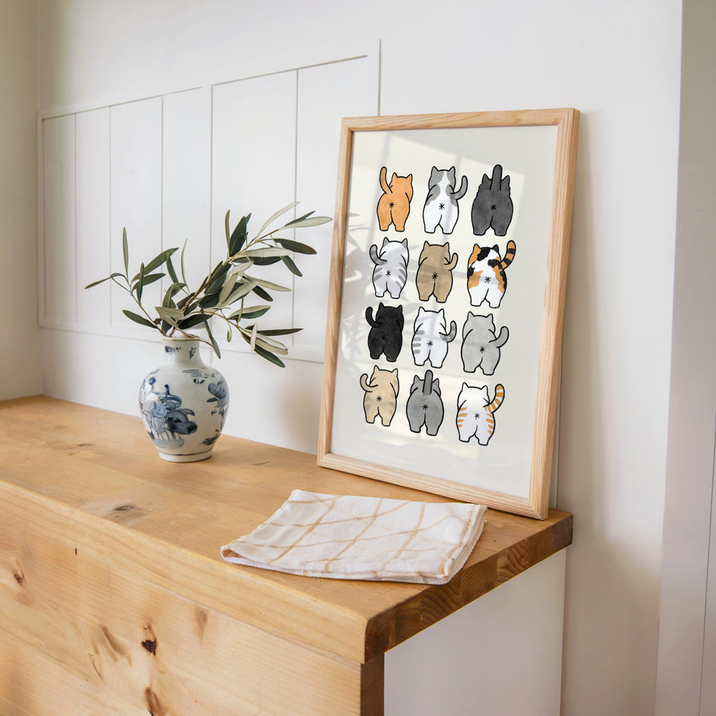 Print leaning on a wooden desk beside a vase and book, simple linework cat butts, ideal cat lover gag gift and bathroom wall art