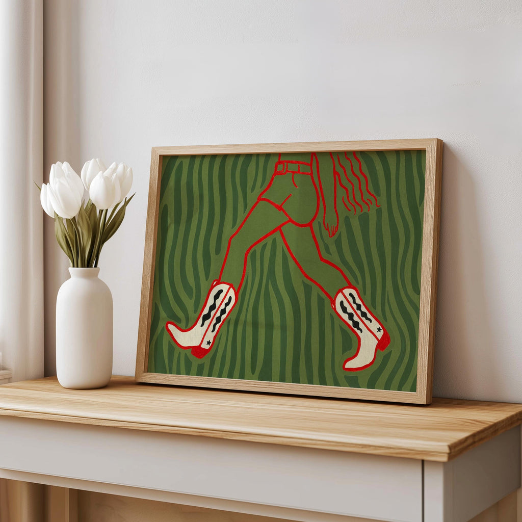 Green patterned cowgirl wall art print leaning on shelf with vase, bold red cowgirl legs and cowboy boots visible