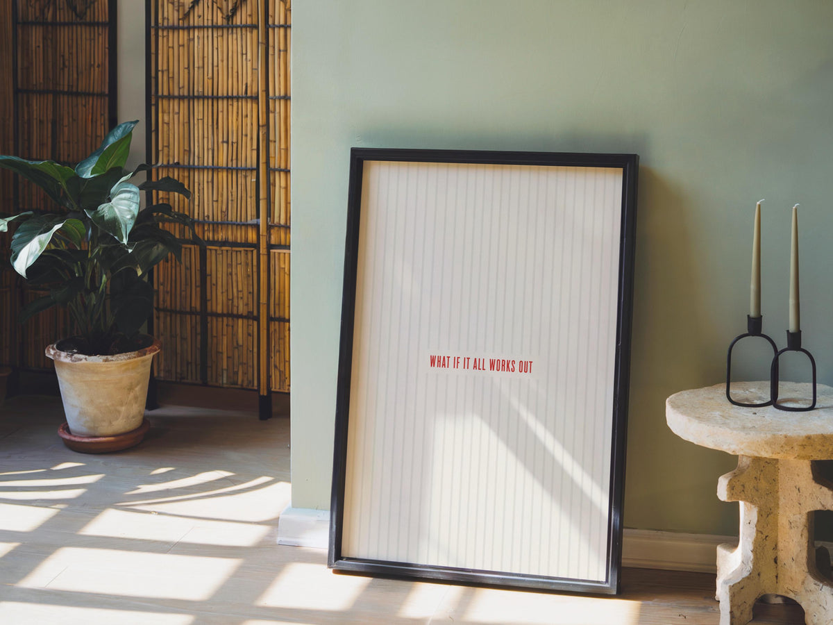Framed what if it all works out poster leaning in a sunlit corner, potted plant and stool