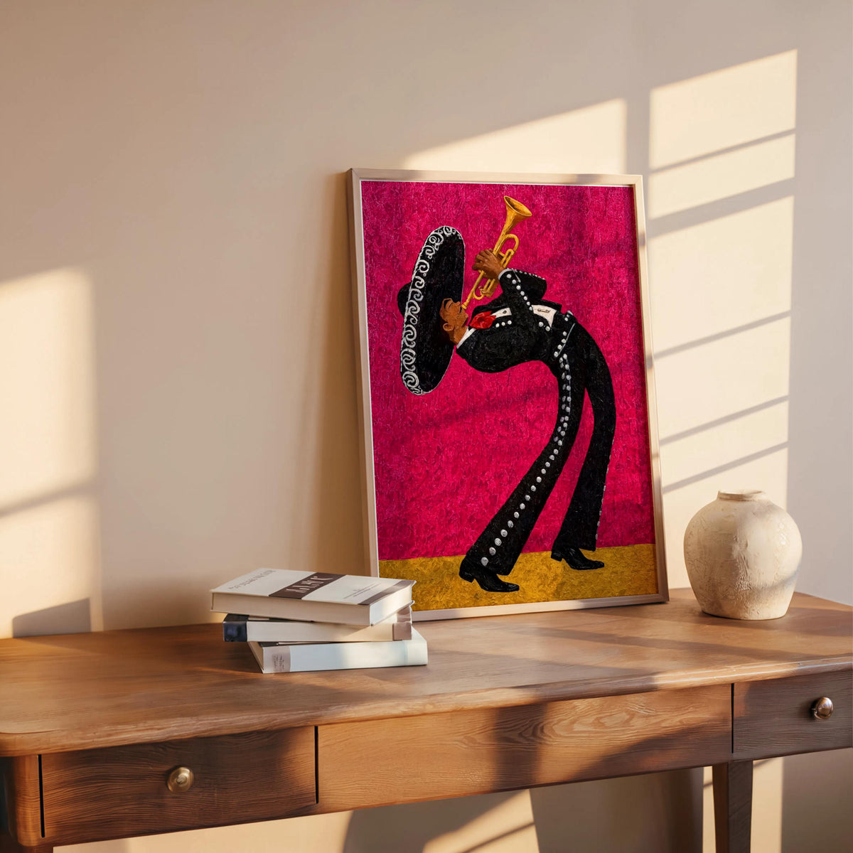 Bright magenta mariachi wall art print leaning on wooden table, vase and stacked books beside it, warm afternoon light