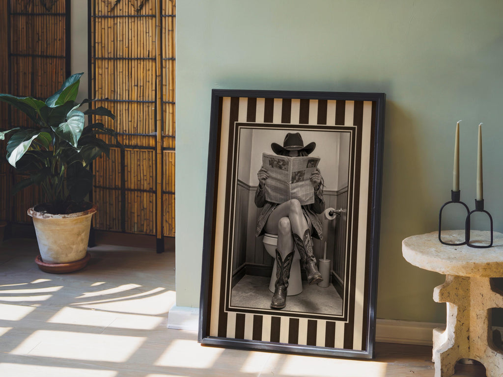 Leaning framed sepia cowgirl print on floor near potted palm, rustic light highlights striped western border, funny cowgirl bathroom wall art