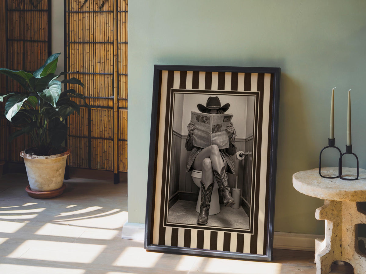 Leaning framed sepia cowgirl print on floor near potted palm, rustic light highlights striped western border, funny cowgirl bathroom wall art