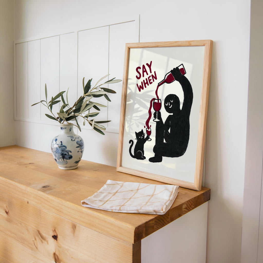 Black SAY WHEN print leaning on desk next to a ceramic vase, warm natural light, wine enthusiast gift, funny cat wine wall art