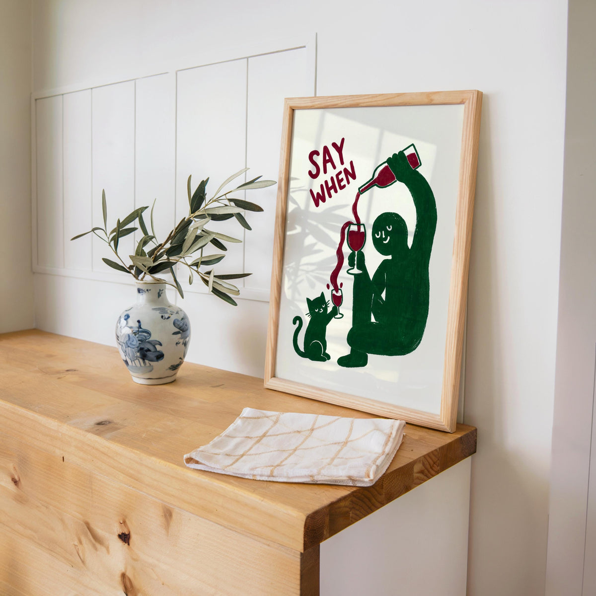 Green SAY WHEN print leaning on wooden desk next to blue vase and greens, retro wine poster, funny cat wine wall art