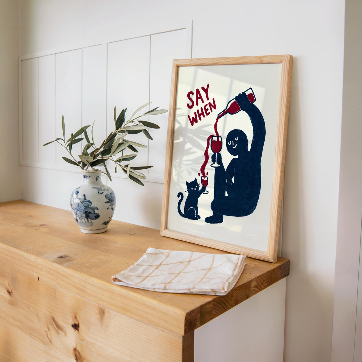 Navy SAY WHEN print propped on a wooden desk beside a ceramic vase and paper, cat wine print, funny cat wine wall art