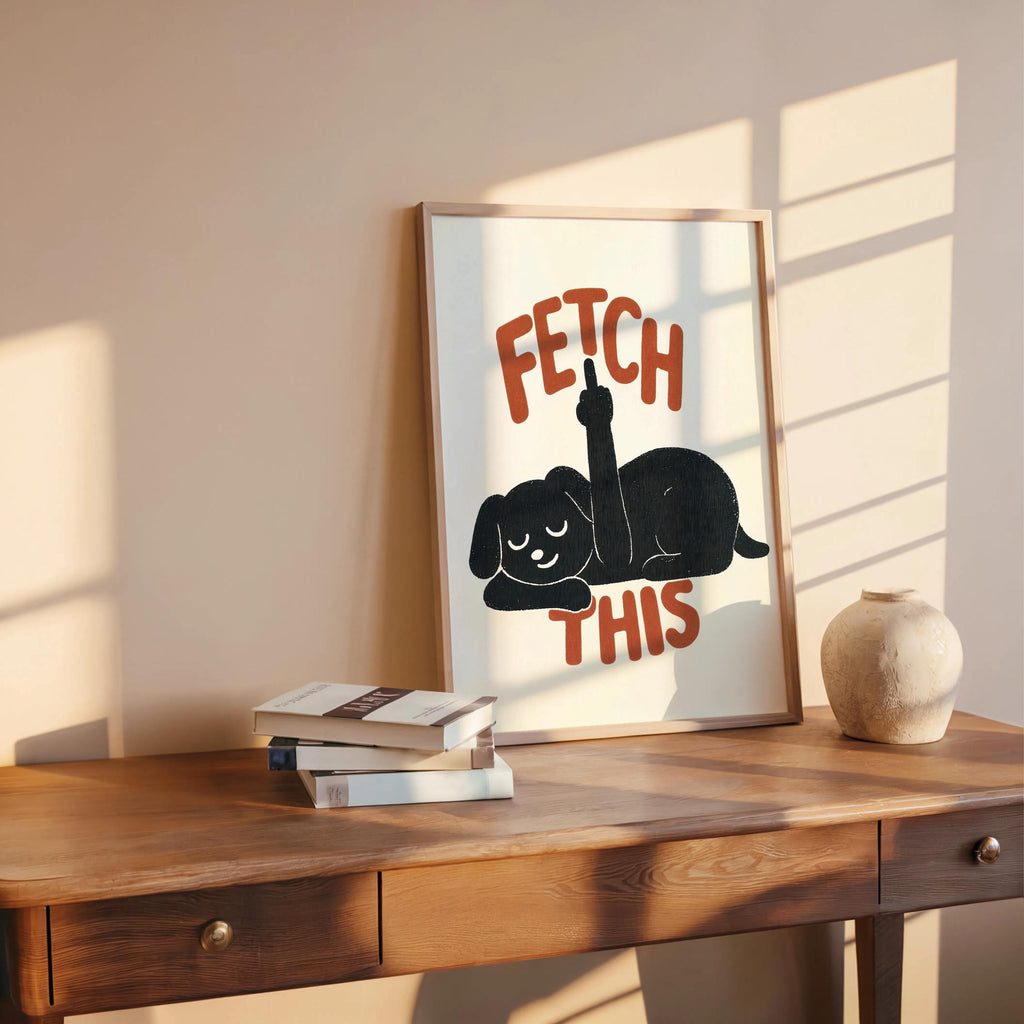 Framed poster casually leaning on a desk with warm sunlight, charcoal dog silhouette and orange text, playful dog decor