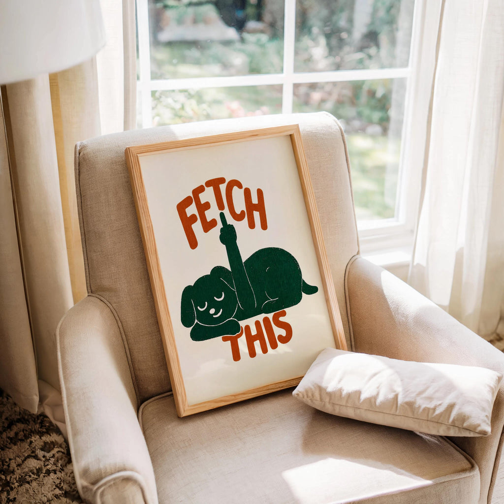 Framed print tucked into a beige armchair by a window, green dog silhouette with orange FETCH THIS lettering, cheeky dog print