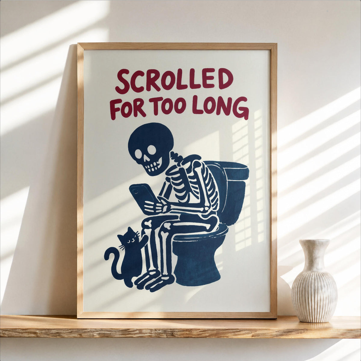 Framed vintage-style funny toilet wall art leaning on shelf, navy skeleton scrolling, cream background, curious cat