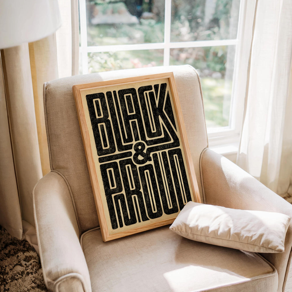 Black and Proud wall art leaning on an upholstered armchair by the window, bold black typography on aged cream paper