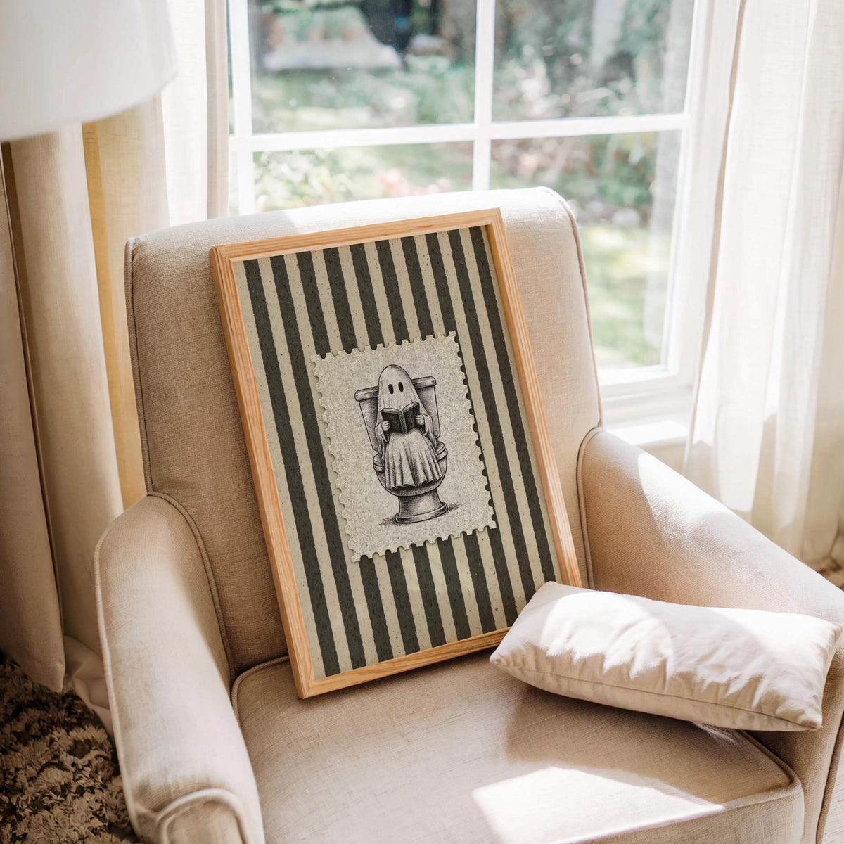 Framed vintage-style ghost toilet print resting on an upholstered armchair by a window, muted cream and olive tones, funny toilet wall art and funny bathroom wall art