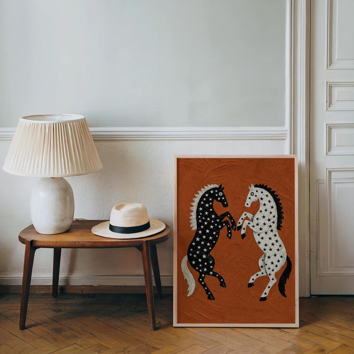 Leaning western horse print beside a lamp and hat on a wooden stool, cozy floor setting, bold black and white rearing horses on rust orange