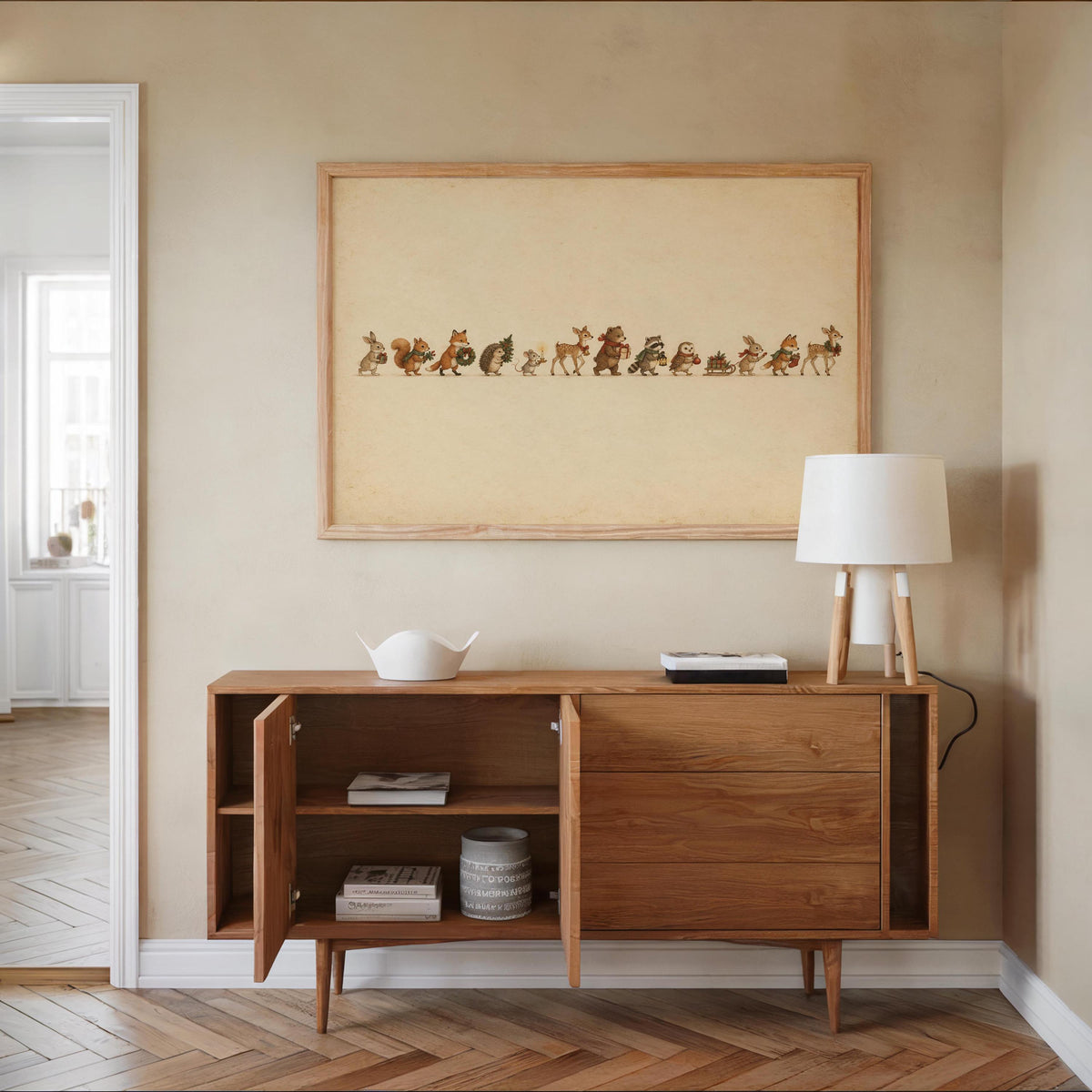 Woodland animal Christmas print centered above a wooden sideboard, lamp, bowl, warm neutral decor