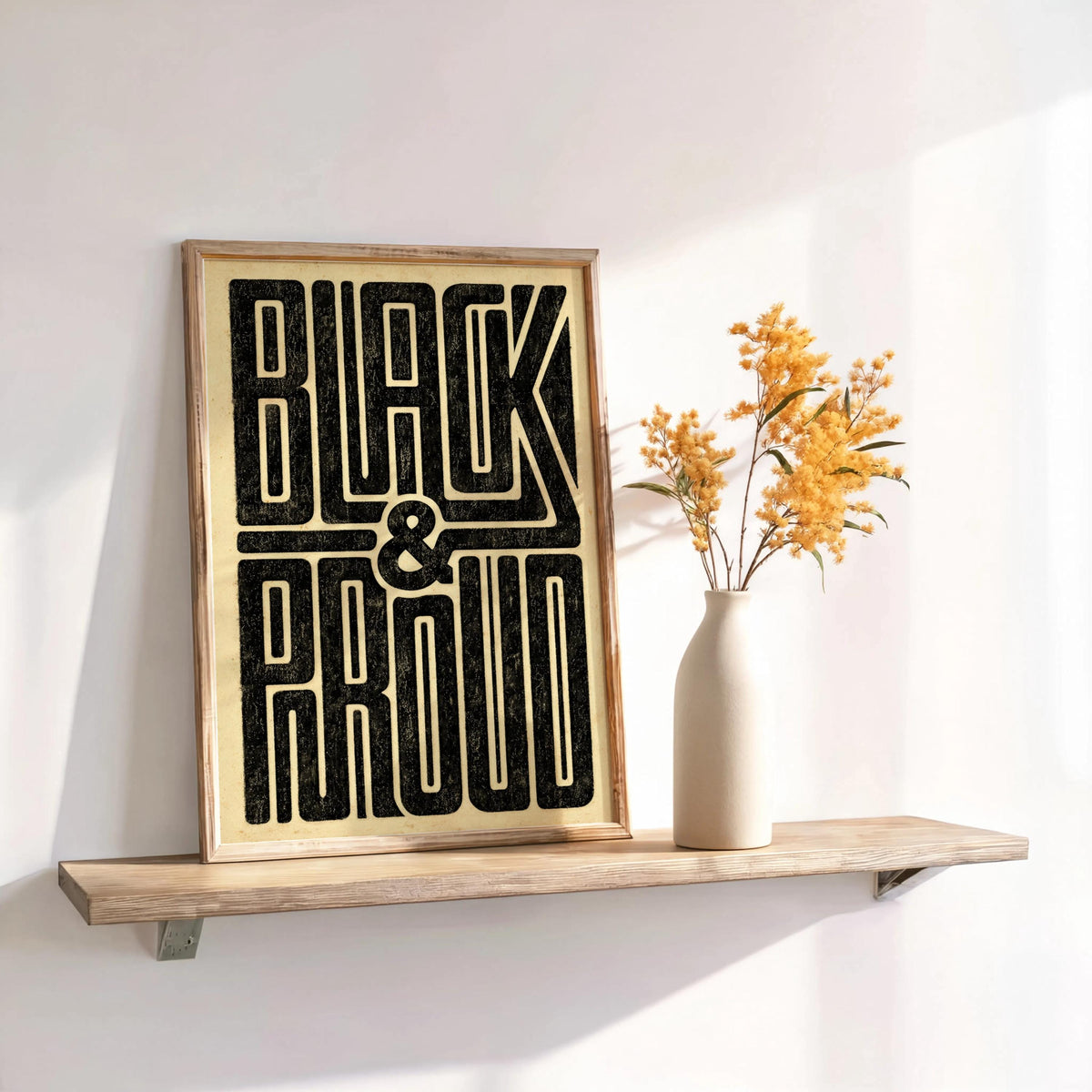 Black and Proud wall art leaning on a narrow shelf with a vase of dried flowers, bold retro black typography