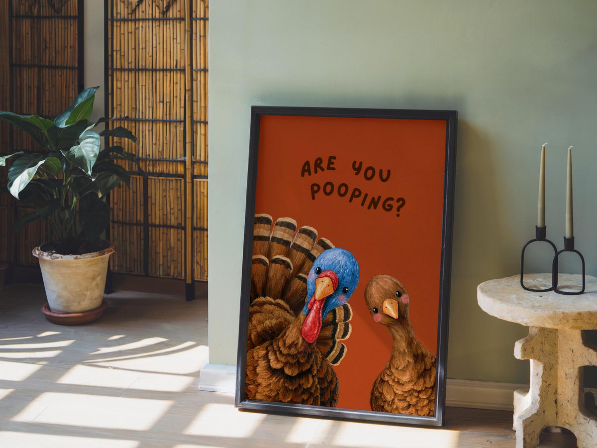 Styled vignette showing funny turkey bathroom print with burnt orange background, leaning by plant for quirky toilet sign display