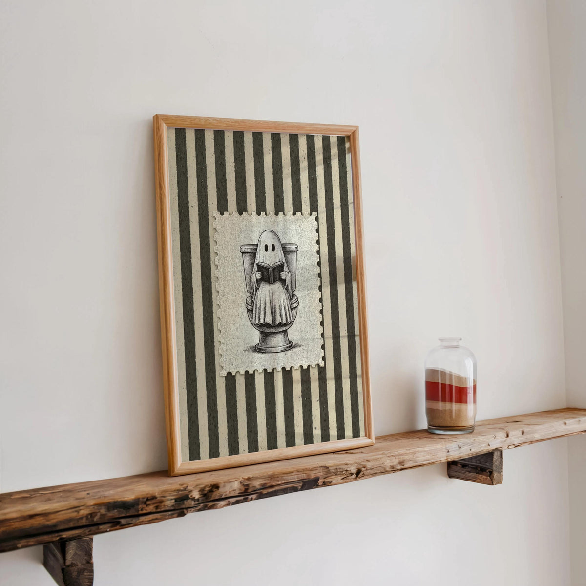 Framed striped Halloween print sitting on a narrow ledge beside a candle jar, distressed border and charcoal tones, funny bathroom wall art
