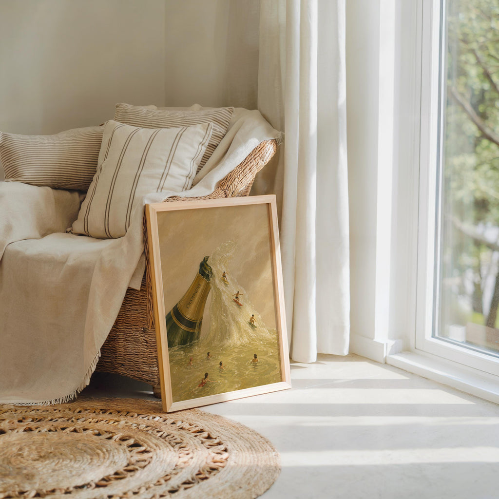 Two champagne bottle wall art prints casually stacked by a comfy chair, neutral textiles, miniature world vibe