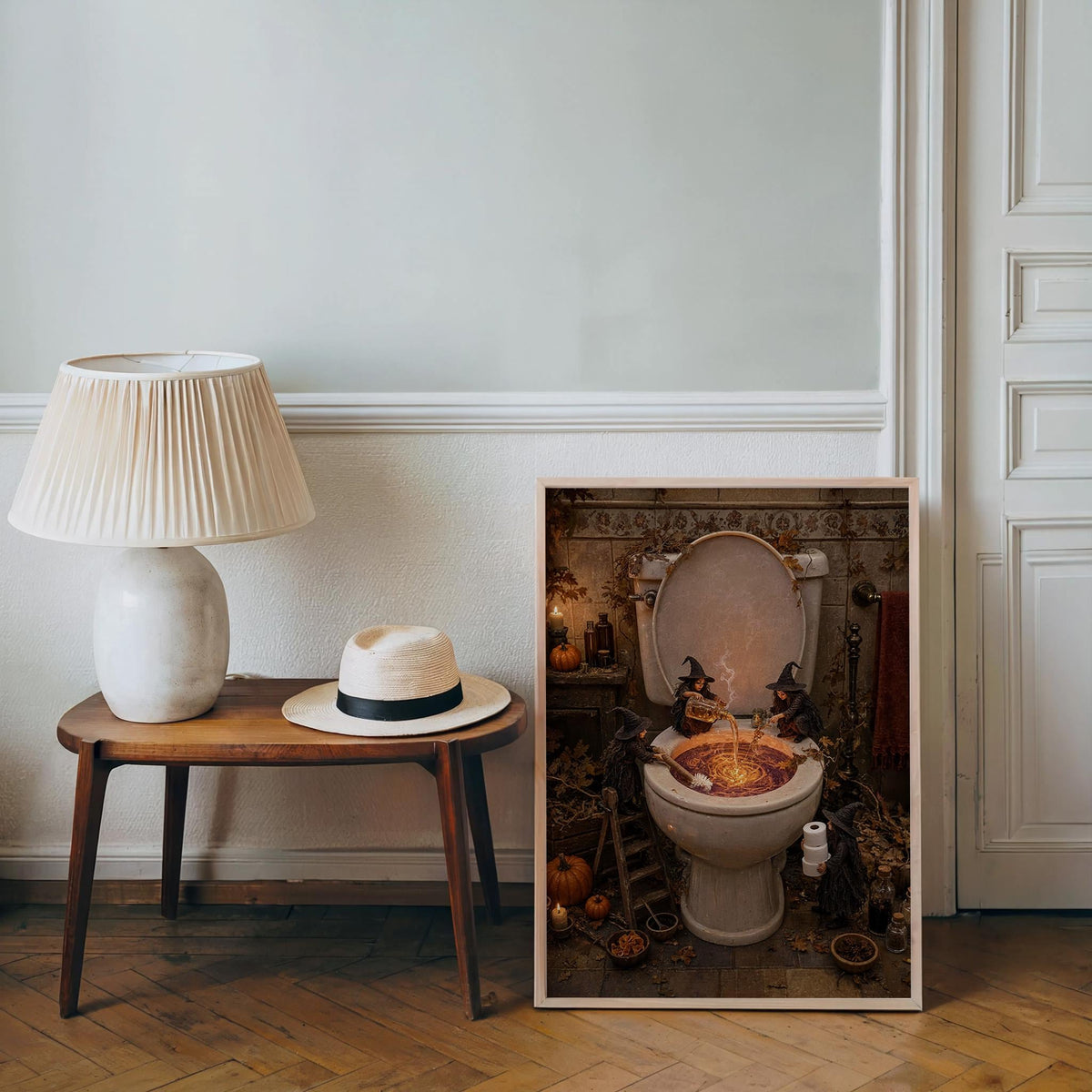 Leaning spooky toilet wall art beside a midcentury table lamp and straw hat, gothic bathroom print charm.