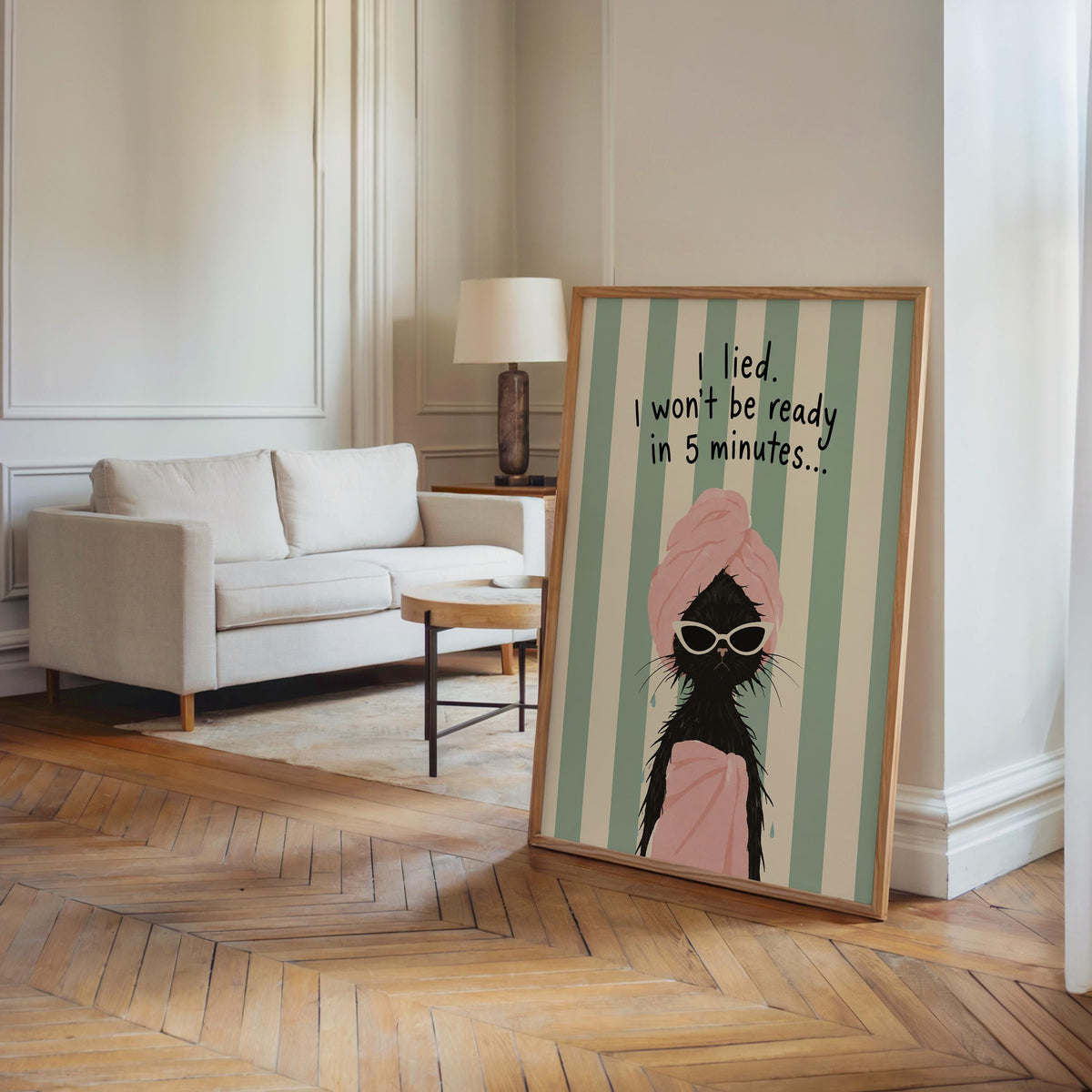 Framed quirky bathroom poster angled on hardwood floor beside sofa, mint stripes and pink towel cat