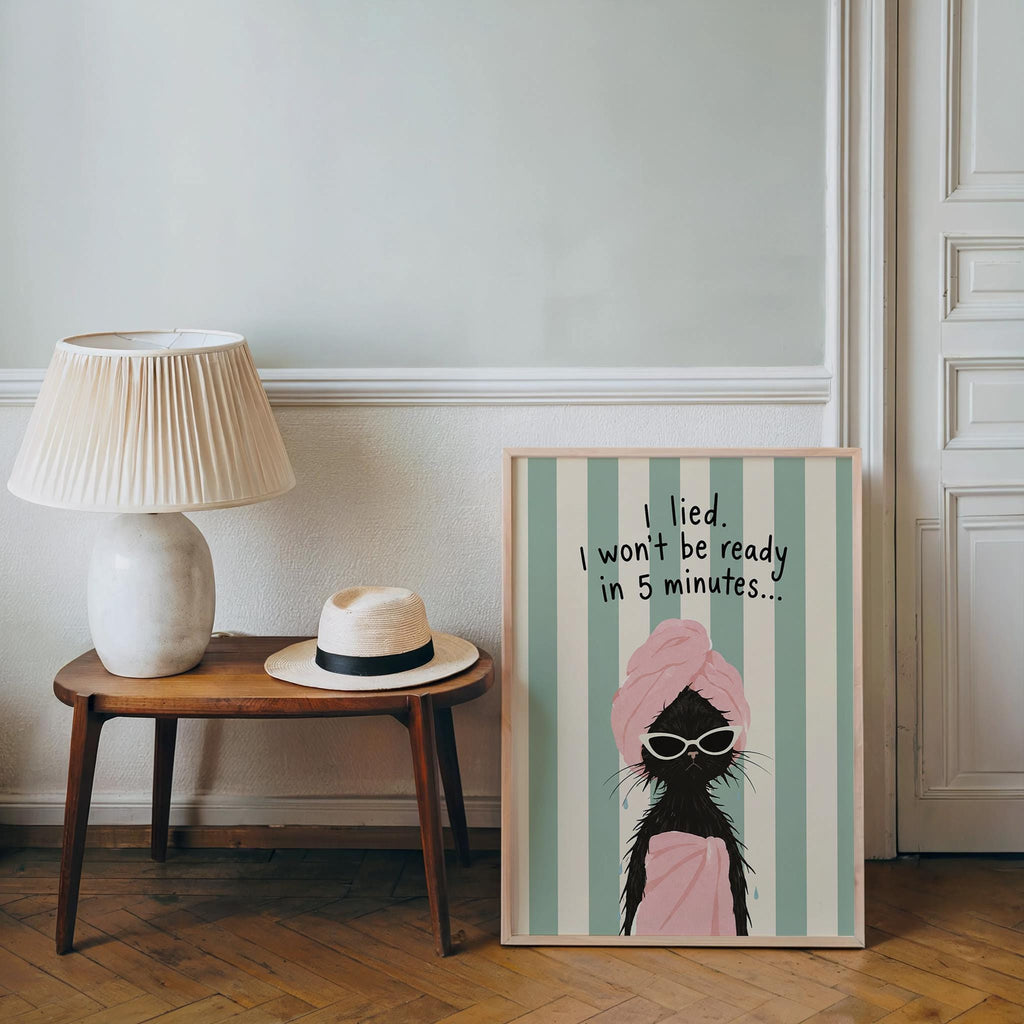 Framed funny black cat bathroom print placed on floor by side table and lamp, cozy neutral room styling