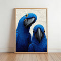 Framed vertical blue macaw wall art leaning on hardwood floor, cobalt macaws with yellow-rimmed eyes